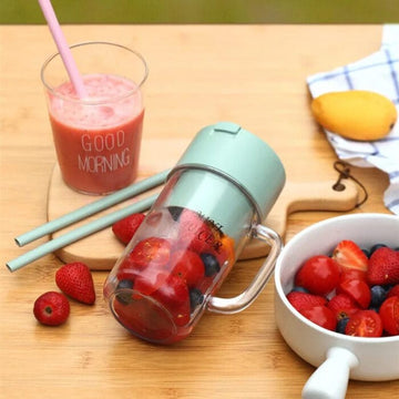 Green tumbler with fruit, glass of juice, and bowl of berries on a wooden table.