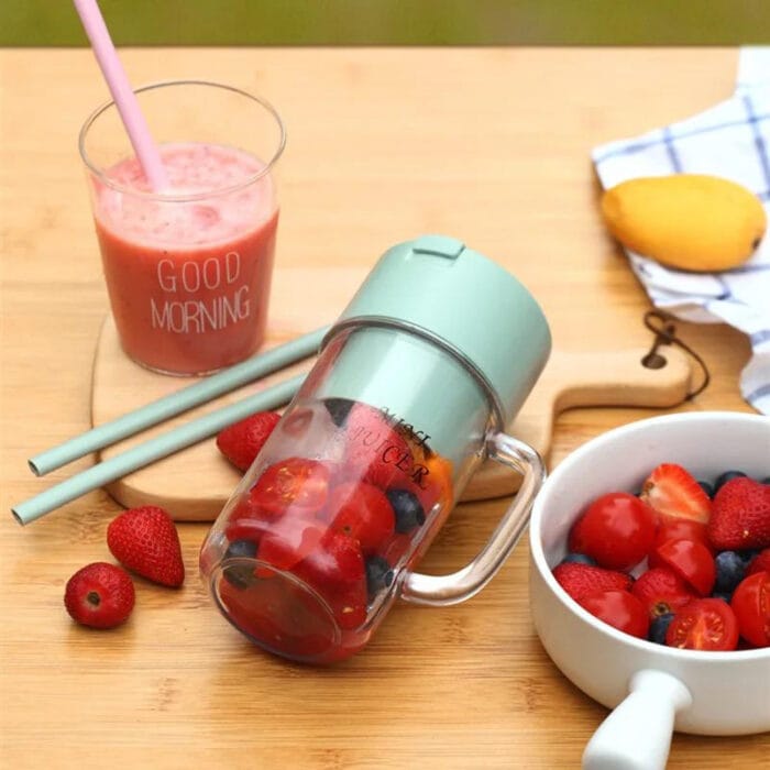 Green tumbler with fruit, glass of juice, and bowl of berries on a wooden table.
