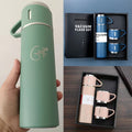 Green vacuum flask held by a hand, with pink flask set in a box on a wooden surface.